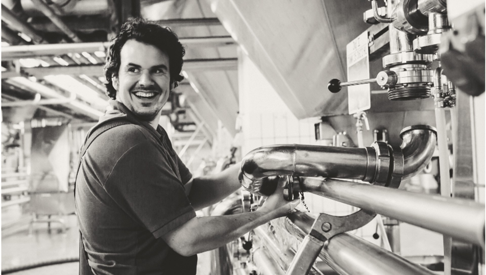 A smiling man adjusts a large metal pipe in an industrial setting.