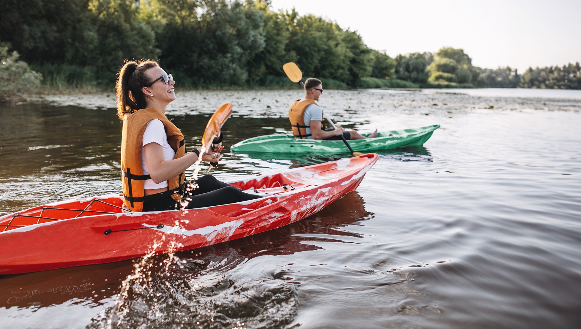 A man and a woman paddle kayaks on a river, wearing life jackets and smiling under the sunlight.