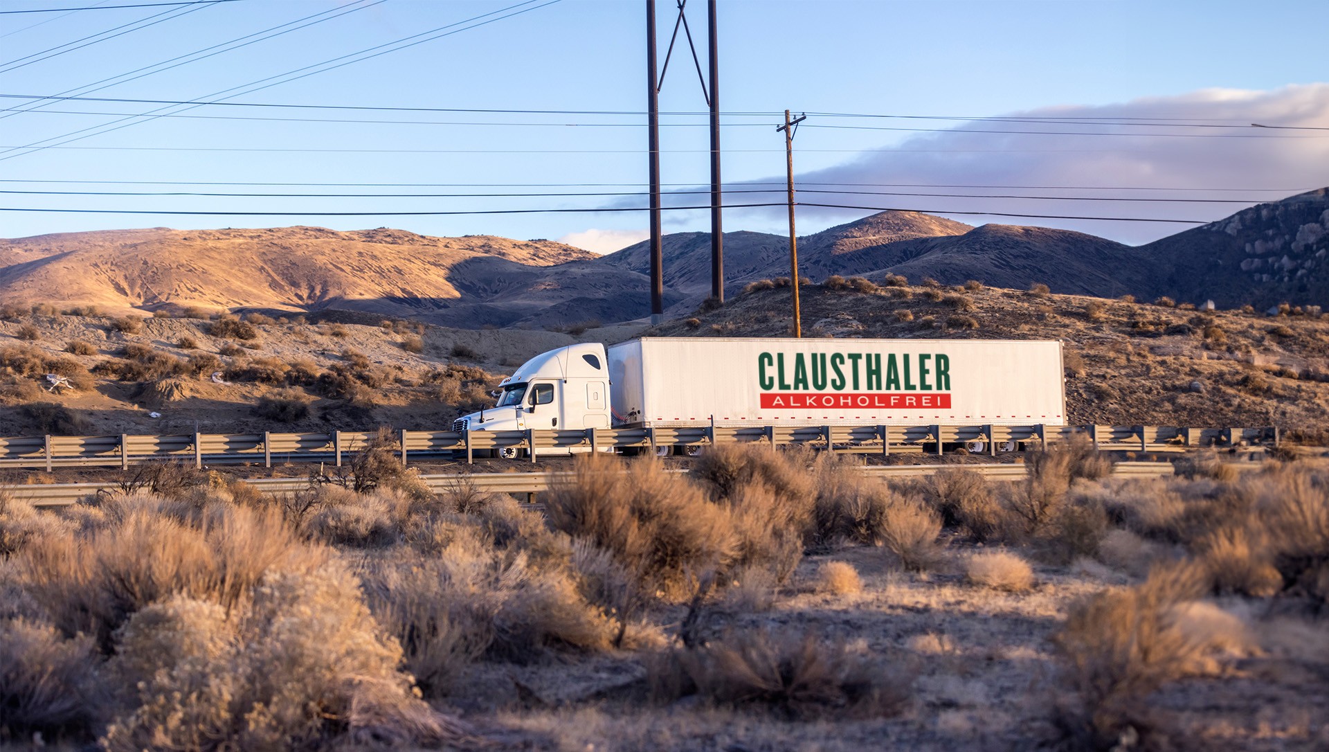A white semi truck hauls a trailer along a highway bordered by arid landscape and hills at dusk.