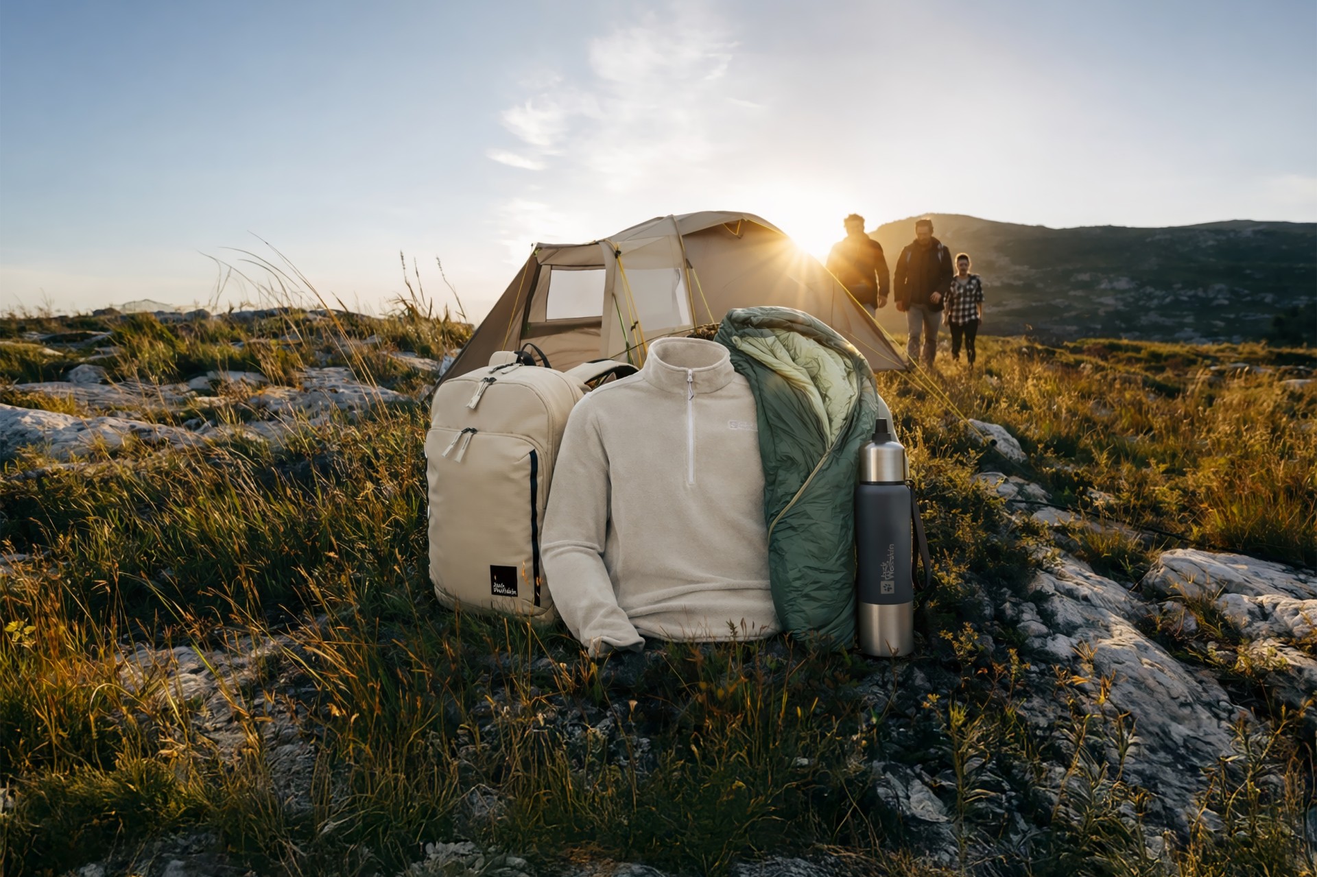 Ein Zelt, eine Tragetasche, eine Jacke, ein Schlafsack und eine Thermosflasche sind auf einem felsigen Hügel im Sonnenuntergang angeordnet, drei Personen befinden sich im Hintergrund.