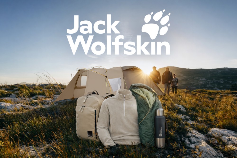 Ein Zelt, eine Tragetasche, eine Jacke, ein Schlafsack und eine Thermosflasche sind auf einem felsigen Hügel im Sonnenuntergang angeordnet, drei Personen befinden sich im Hintergrund.